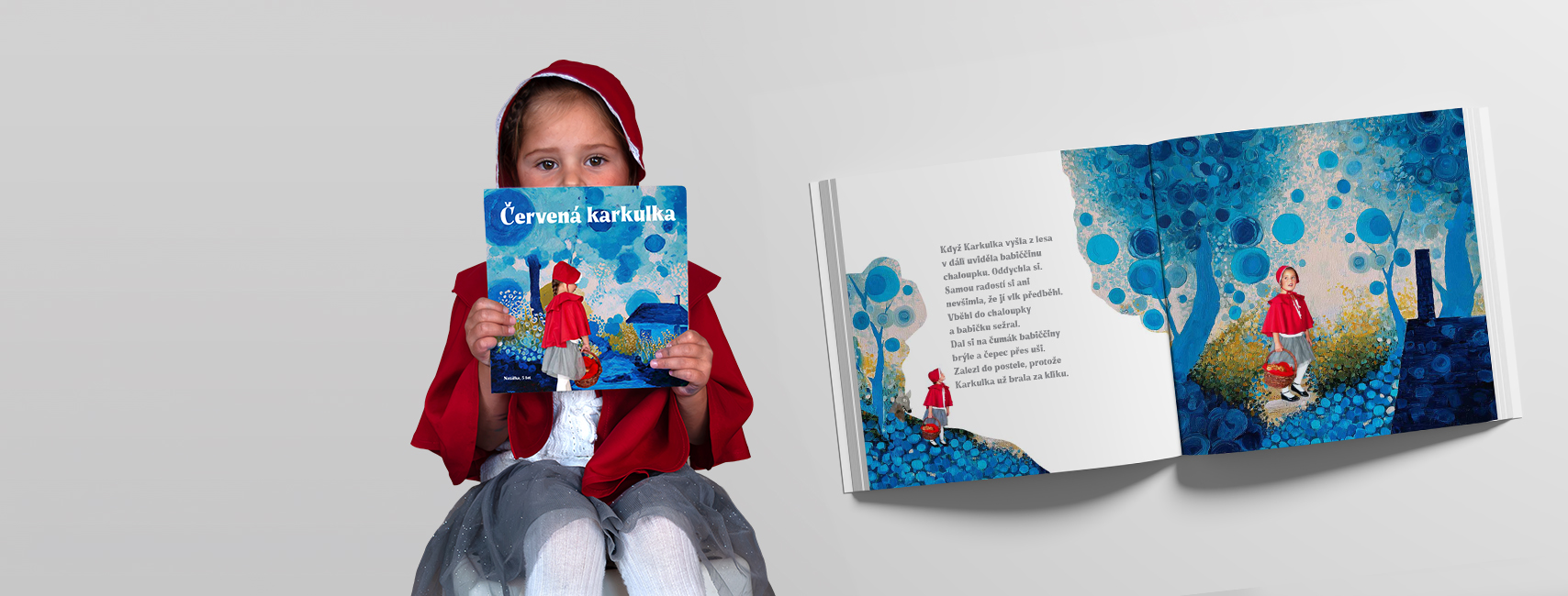 Holčička v červené pelerínce drží knihu Červená karkulka, vedle rozevřené album s modrými malovanými ilustracemi
